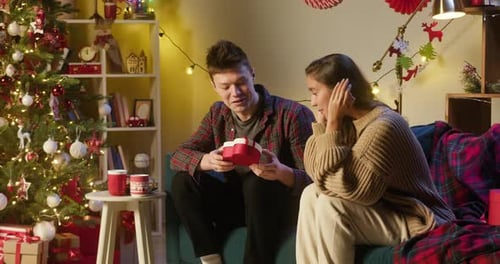 Young Man Giving Present Box to His Girlfriend Sitting on Sofa in Living Room