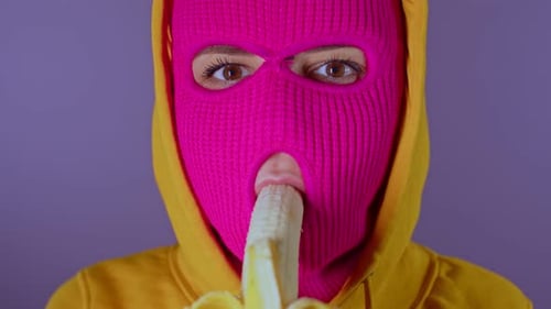 Close up of young woman in pink balaclava and yellow hood eats banana on purple background.
