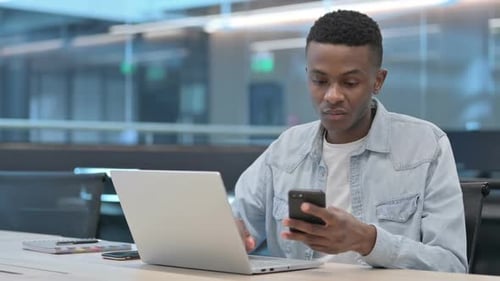 Young Man Using Smartphone at Desk with Laptop
