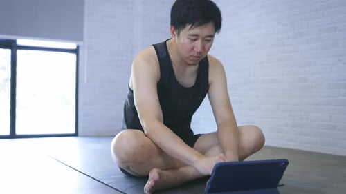 Joven asiático en ropa deportiva haciendo ejercicio en casa mientras ve un vídeo tutorial en línea en una tableta