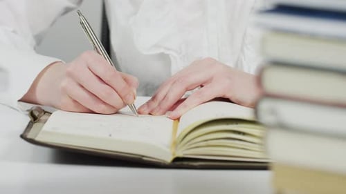Woman Writes in Notebook Beside Stack of Books
