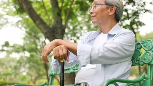Asian senior man holding a cane and sitting on a bench in the park