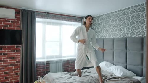 Playful Guy in Bathrobe Dancing on Bed Listening To Music Through Headphones in Hotel
