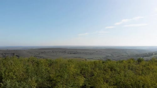 Aerial view over green forest on a hill, revealing a valley with meadows and fields, sunny, summer d