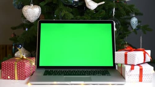 Laptop with Green Screen on Table Among Christmas Gift Boxes and Christmas Tree