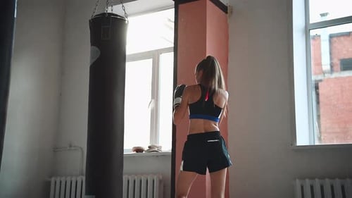 A Girl in Sports Clothes is Engaged in Boxing and Works Out a Punch with Her Hand on a Punching Bag