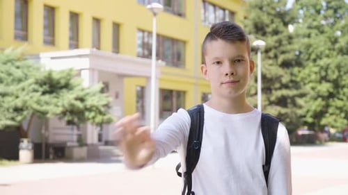 A Caucasian Teenage Boy Waves at the Camera with a Smile a School in the Background
