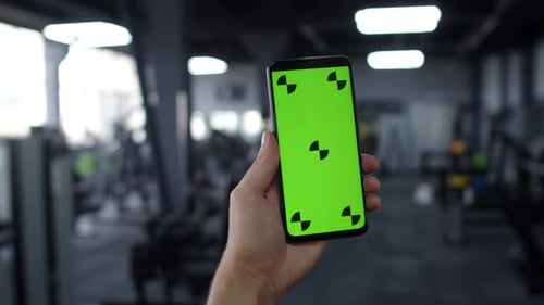 Man's Hand Holds Smartphone with Green Screen on Background of Gym Using Sports App