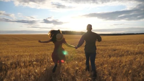 Young Couple Holding Hands of Each Other and Running Through the Wheat Field at Sunset