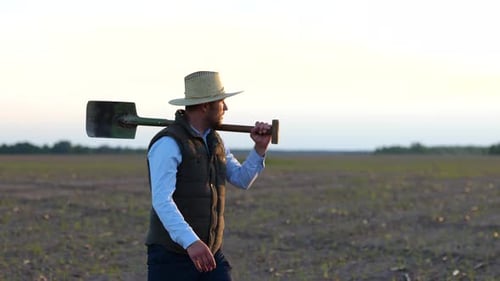 Portrait of Young Caucasian Male Farmer in Hat Walking at Own Farm with Shovel on Shoulder