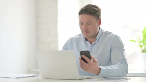 Man Browsing Internet on Smartphone while using Laptop in Office