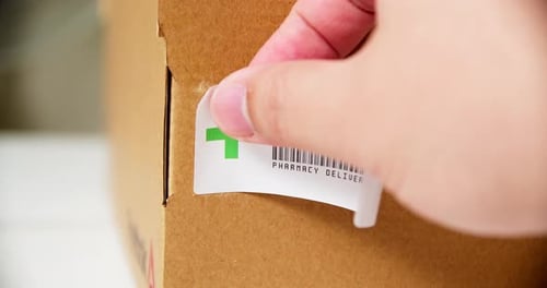 Hände beim Anbringen des +PHARMACY Sticker-Etiketts auf einen Versandkarton