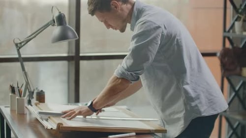 A Man is a Young Modern Architect Sitting at a Table Drawing a Plan of the Building and Developing a