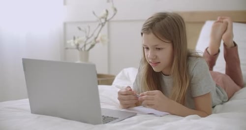 Fröhliche Schülerin antwortet während des Online-Unterrichts per Laptop, Auf dem Bett liegen und sprechen, e.