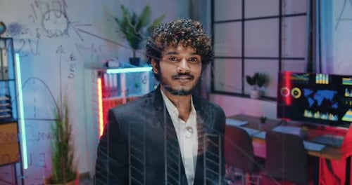 Hombre posando ante la cámara en una oficina nocturna cerca de una pared de cristal mientras pensaba en la estrategia empresarial
