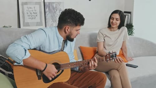 Man Plays Guitar for Smiling Woman on Couch
