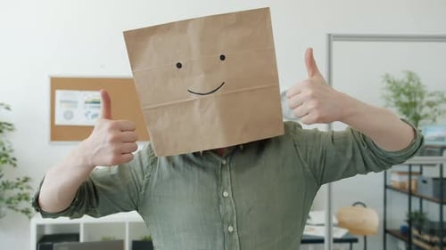 Porträt eines Geschäftsmannes, der Brotsack mit Lächeln-Emoji trägt und am Arbeitsplatz eine Handgeste mit dem Daumen nach oben zeigt