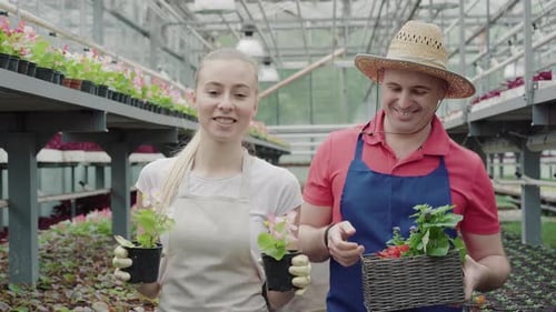 Two Smiling People Holding Flowers and Plants in Greenhouse