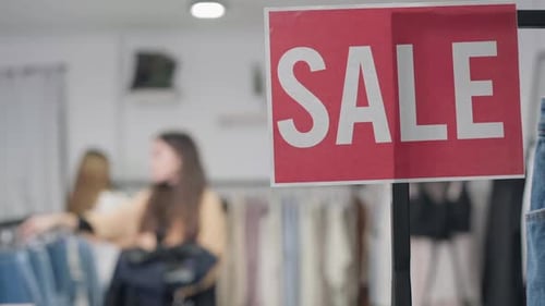 Closeup Sale Message in Clothing Shop with Two Young Women Choosing Apparel at Background Indoors