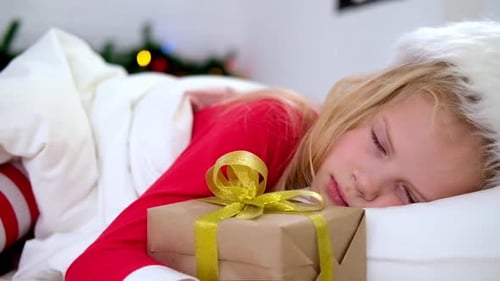 Menina com chapéu de Papai Noel e pijama de Natal deitada na cama com caixa de presente em casa
