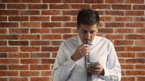 An Eastern-looking Businessman Flips Through a Tape on His Phone and Smokes an E-cigarette. The