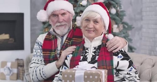 Close-up of Elderly Caucasian Couple Sitting Next To Christmas Tree with Presents, Looking at Camera