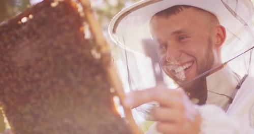 Gesicht eines jungen bärtigen Imkers in weißem Schutzanzug mit Bienenstock-Werkzeug in der Hand, der hält