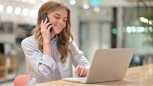 Businesswoman with Laptop Talking on Smartphone in Office