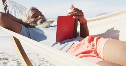 Happy senior african american man reading and lying in hammock on sunny beach