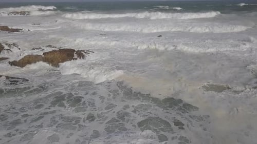 Aerial: Rows of ocean waves break into whitewater on shoreline rocks