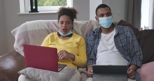Couple With Devices Wearing Masks on Couch