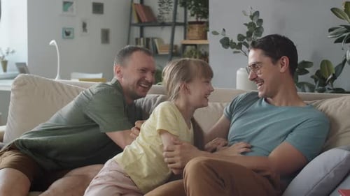 Two Fathers Playfully Tickle Child on Sofa at Home