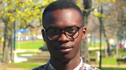 Ein junger schwarzer Mann setzt an einem sonnigen Tag eine Brille auf und lächelt in einem Park in die Kamera - Nahaufnahme