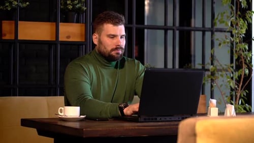 Junger attraktiver bärtiger Mann arbeitet selbstbewusst am Laptop während der Kaffeepause im Café