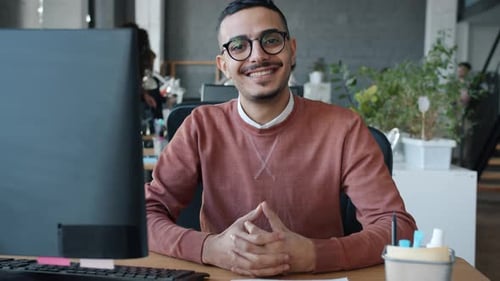 Porträt eines glücklichen arabischen Mannes, der lächelnd am Computertisch im Büro sitzt und in die Kamera blickt