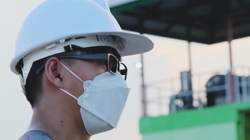 Young Asian engineer wearing a helmet and mask looks and smiles at the camera on the dam background.