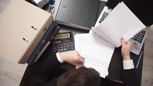 Male office worker is sitting at table with laptop and various stationery on it, looks at working