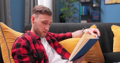 Young Man Reads Book on Couch at Home