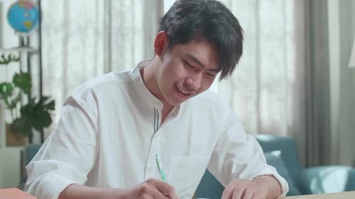 Close Up Of A Happy Asian Man Student Writing In Notebook On The Table While Studying At Home