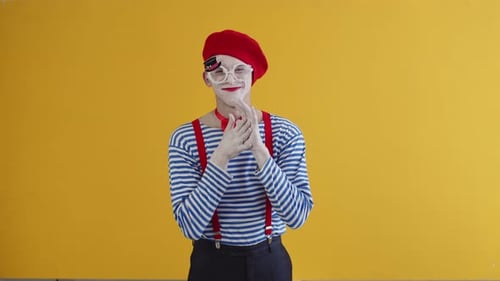 Un mimo codicioso vestido con maquillaje blanco, camisa a rayas y sombrero rojo y tirantes frotándose las manos