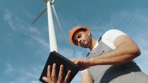 Indischer Ingenieur in Helm und grauem Overall steht zwischen Windmühlenfarm mit