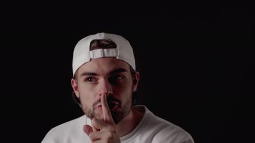 Trendy 20s Man shushing into the camera, silence gesture, close, black background