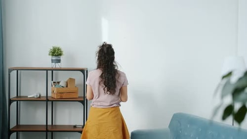 Back View of Young Woman Walking to Wall and Hanging Modern Picture After Relocation