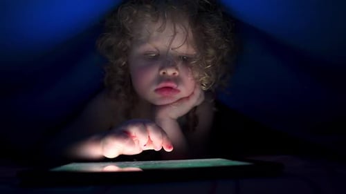 La nuit, une petite fille caucasienne utilise une tablette sous une couverture sur le lit avant de se coucher.