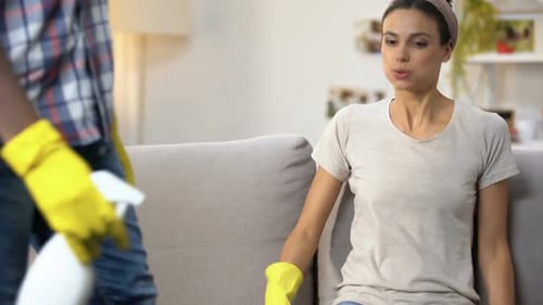 Pareja de raza mixta cansada en guantes con spray limpiador sentado en el sofá, tareas domésticas