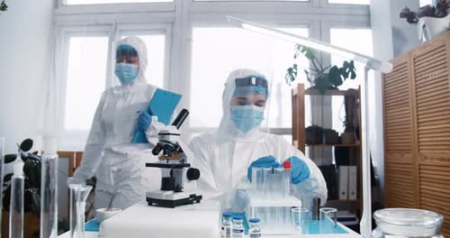 Fokussierte junge Laborwissenschaftlerin im vollständigen medizinischen Schutzanzug und Schild arbeitet an neuen