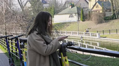 Woman Uses Phone Near Electric Scooters in City Park