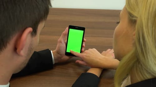 Two Office Workers, Man and Woman, Sit at A Desk and Slide at A Smartphone with A Green Screen