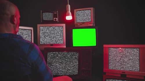 Un homme est assis devant un groupe de 9 téléviseurs vintage avec écrans Chroma Key dans une pièce au mur noir