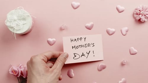 Hand Placing Mother's Day Card Among Gifts and Flowers
